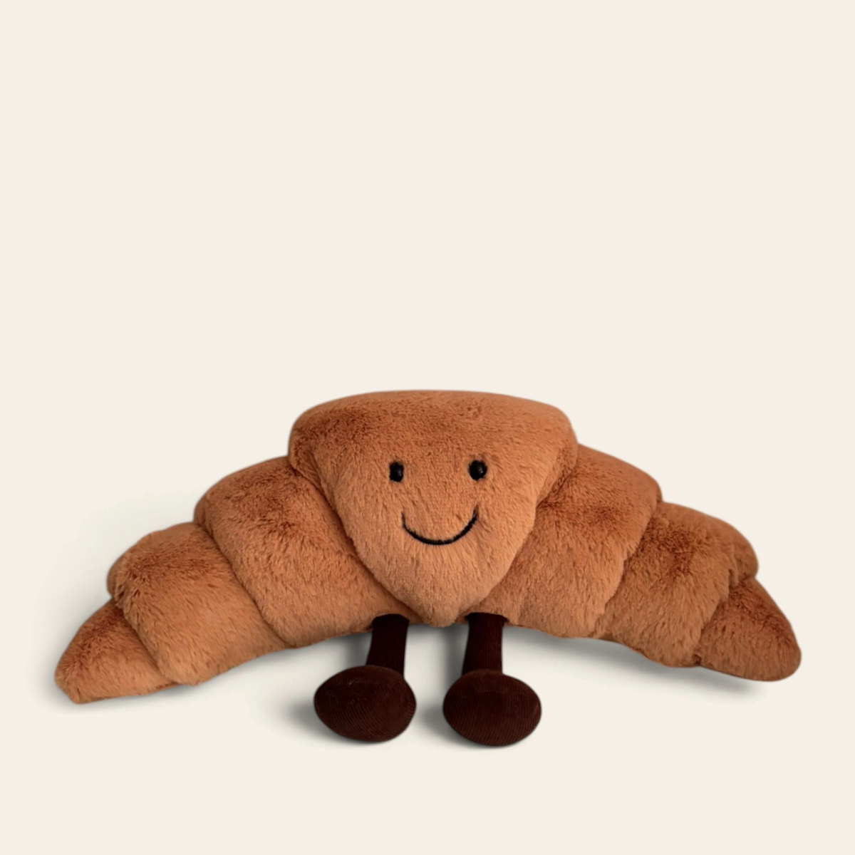 Amuseables Croissant A2CRO: a golden brown plush croissant toy with a smiling face and dark brown legs, lying flat