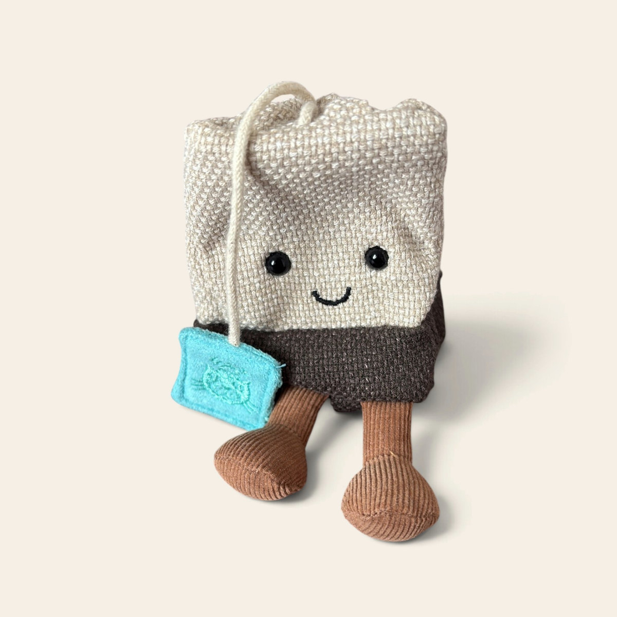 Amuseables Steepy Tea Bag A4TEAB: a plush tea bag toy with a cream woven top, a dark brown base, a string with a blue tag, a smiling face and brown corduroy legs
